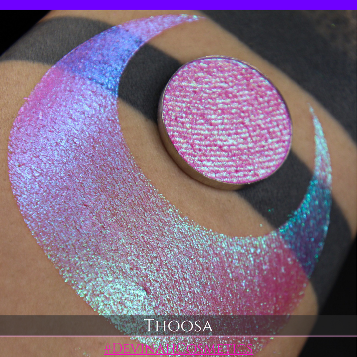 A round eyeshadow pan labeled 'THOOSA' made by Devinah Cosmetics, placed over a large crescent moon swatch of the same shimmering shifty eyeshadow on skin. The eyeshadow has a duochrome multichrome shift between colors blue, purple, green, pink.