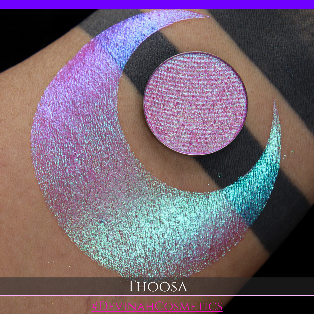 A round eyeshadow pan labeled 'THOOSA' made by Devinah Cosmetics, placed over a large crescent moon swatch of the same shimmering shifty eyeshadow on skin. The eyeshadow has a duochrome multichrome shift between colors blue, purple, green, pink.