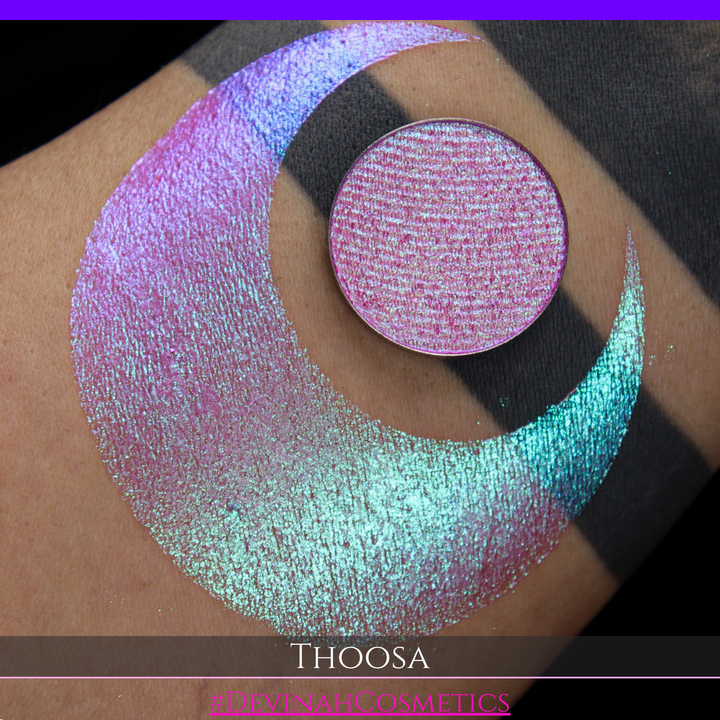 A round eyeshadow pan labeled 'THOOSA' made by Devinah Cosmetics, placed over a large crescent moon swatch of the same shimmering shifty eyeshadow on skin. The eyeshadow has a duochrome multichrome shift between colors blue, purple, green, pink.