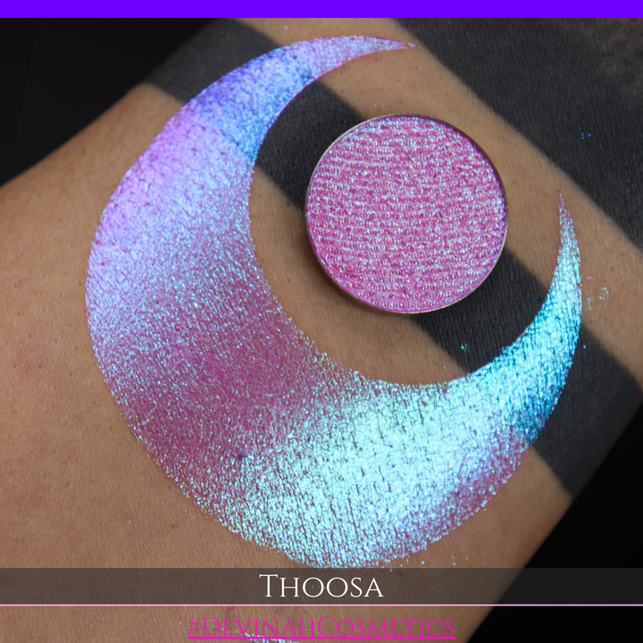 A round eyeshadow pan labeled 'THOOSA' made by Devinah Cosmetics, placed over a large crescent moon swatch of the same shimmering shifty eyeshadow on skin. The eyeshadow has a duochrome multichrome shift between colors blue, purple, green, pink.