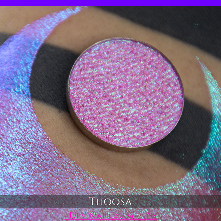 A round eyeshadow pan labeled 'THOOSA' made by Devinah Cosmetics, placed over a large crescent moon swatch of the same shimmering shifty eyeshadow on skin. The eyeshadow has a duochrome multichrome shift between colors blue, purple, green, pink.