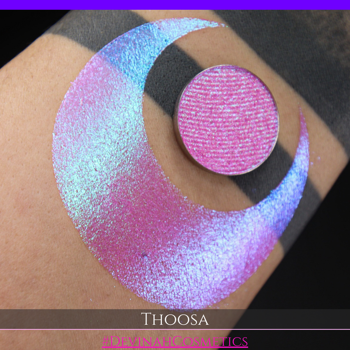 A round eyeshadow pan labeled 'THOOSA' made by Devinah Cosmetics, placed over a large crescent moon swatch of the same shimmering shifty eyeshadow on skin. The eyeshadow has a duochrome multichrome shift between colors blue, purple, green, pink.