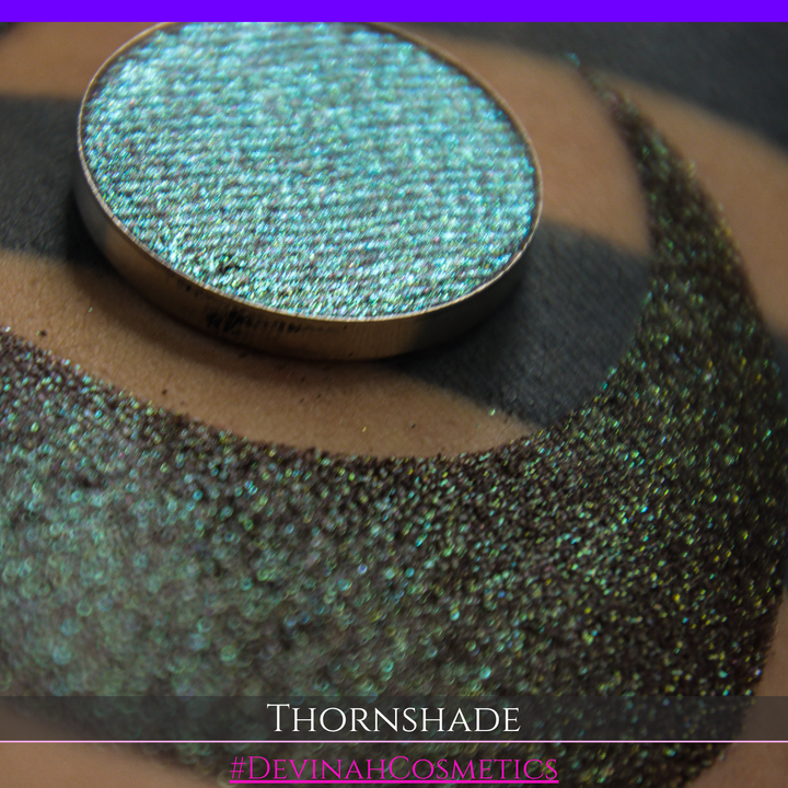 A close-up of a person's arm with a large crescent-shaped swatch of shimmering brown to green eyeshadow, alongside a round eyeshadow pan showing the product texture. Text at the bottom reads 'THORNSHADE' and '#DEVINAHCOSMETICS'.
