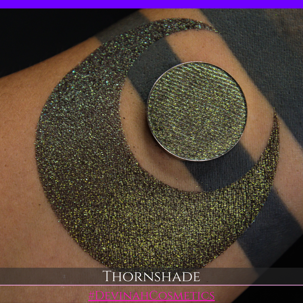 A close-up of a person's arm with a large crescent-shaped swatch of shimmering brown to green eyeshadow, alongside a round eyeshadow pan showing the product texture. Text at the bottom reads 'THORNSHADE' and '#DEVINAHCOSMETICS'.