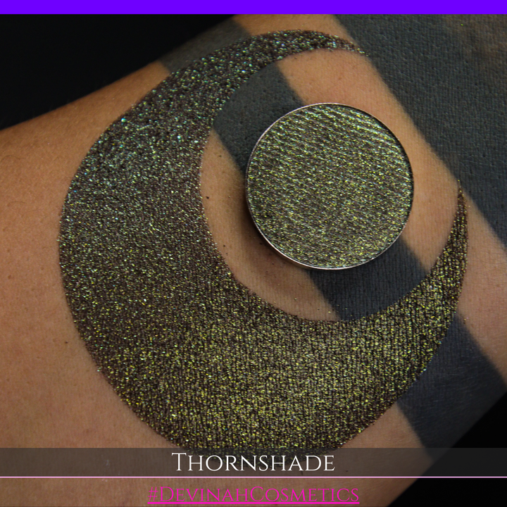 A close-up of a person's arm with a large crescent-shaped swatch of shimmering brown to green eyeshadow, alongside a round eyeshadow pan showing the product texture. Text at the bottom reads 'THORNSHADE' and '#DEVINAHCOSMETICS'.