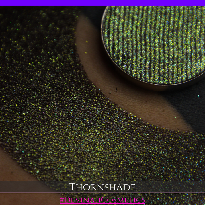 A close-up of a person's arm with a large crescent-shaped swatch of shimmering brown to green eyeshadow, alongside a round eyeshadow pan showing the product texture. Text at the bottom reads 'THORNSHADE' and '#DEVINAHCOSMETICS'.