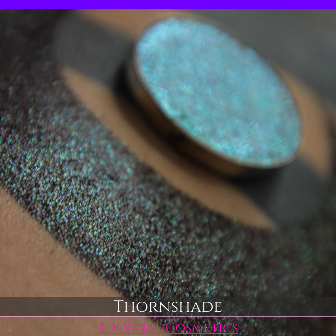 A close-up of a person's arm with a large crescent-shaped swatch of shimmering brown to green eyeshadow, alongside a round eyeshadow pan showing the product texture. Text at the bottom reads 'THORNSHADE' and '#DEVINAHCOSMETICS'.