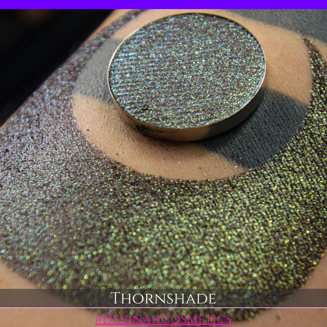 A close-up of a person's arm with a large crescent-shaped swatch of shimmering brown to green eyeshadow, alongside a round eyeshadow pan showing the product texture. Text at the bottom reads 'THORNSHADE' and '#DEVINAHCOSMETICS'.