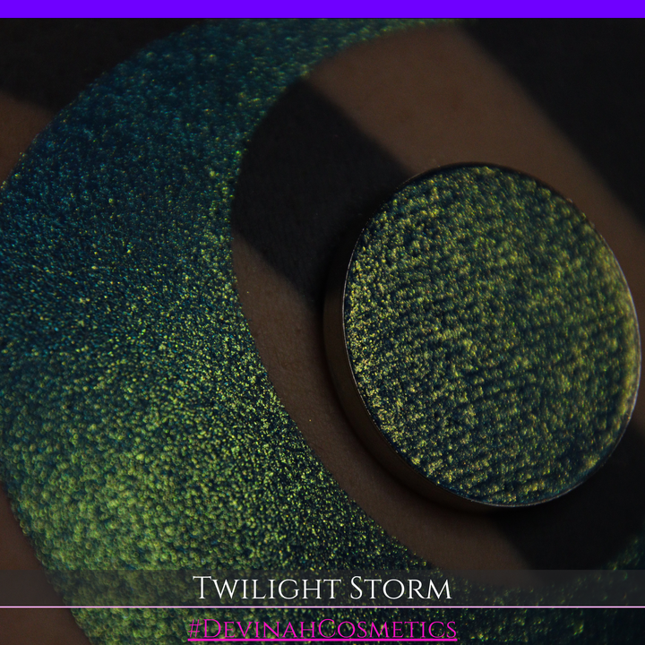 A close-up of a person's arm with a large crescent-shaped eyeshadow swatch in a shimmering teal-to-lime green gradient, with a round pan of the same eyeshadow placed above it. Text at the bottom reads 'TWILIGHT STORM' and '#DEVINAHCOSMETICS'.