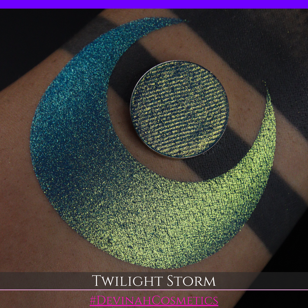 A close-up of a person's arm with a large crescent-shaped eyeshadow swatch in a shimmering teal-to-lime green gradient, with a round pan of the same eyeshadow placed above it. Text at the bottom reads 'TWILIGHT STORM' and '#DEVINAHCOSMETICS'.