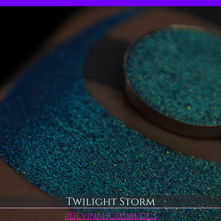A close-up of a person's arm with a large crescent-shaped eyeshadow swatch in a shimmering teal-to-lime green gradient, with a round pan of the same eyeshadow placed above it. Text at the bottom reads 'TWILIGHT STORM' and '#DEVINAHCOSMETICS'.