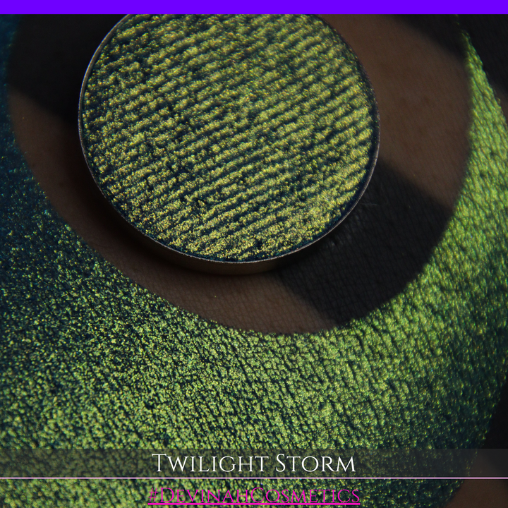 A close-up of a person's arm with a large crescent-shaped eyeshadow swatch in a shimmering teal-to-lime green gradient, with a round pan of the same eyeshadow placed above it. Text at the bottom reads 'TWILIGHT STORM' and '#DEVINAHCOSMETICS'.