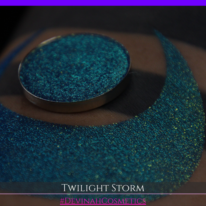 A close-up of a person's arm with a large crescent-shaped eyeshadow swatch in a shimmering teal-to-lime green gradient, with a round pan of the same eyeshadow placed above it. Text at the bottom reads 'TWILIGHT STORM' and '#DEVINAHCOSMETICS'.