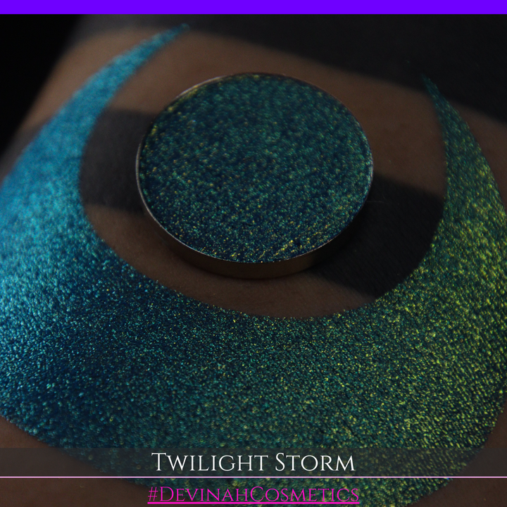 A close-up of a person's arm with a large crescent-shaped eyeshadow swatch in a shimmering teal-to-lime green gradient, with a round pan of the same eyeshadow placed above it. Text at the bottom reads 'TWILIGHT STORM' and '#DEVINAHCOSMETICS'.