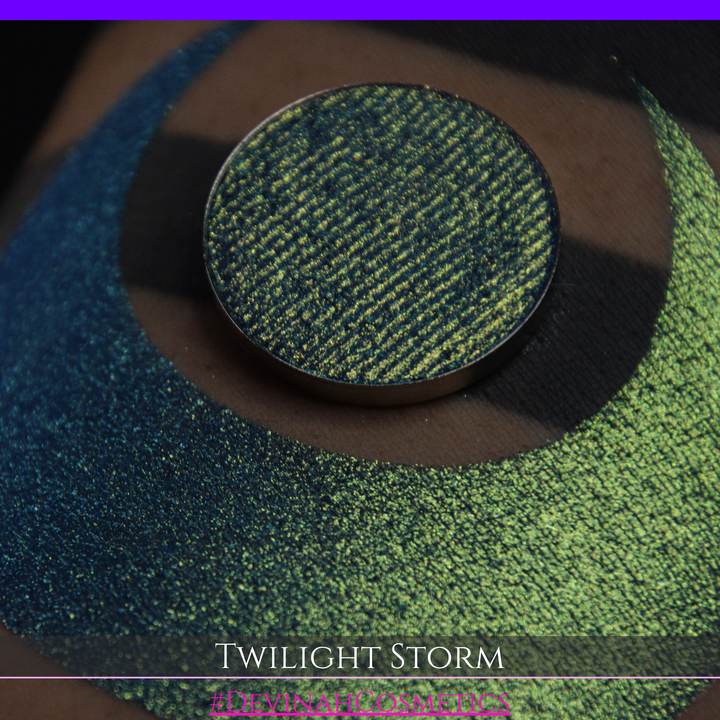 A close-up of a person's arm with a large crescent-shaped eyeshadow swatch in a shimmering teal-to-lime green gradient, with a round pan of the same eyeshadow placed above it. Text at the bottom reads 'TWILIGHT STORM' and '#DEVINAHCOSMETICS'.