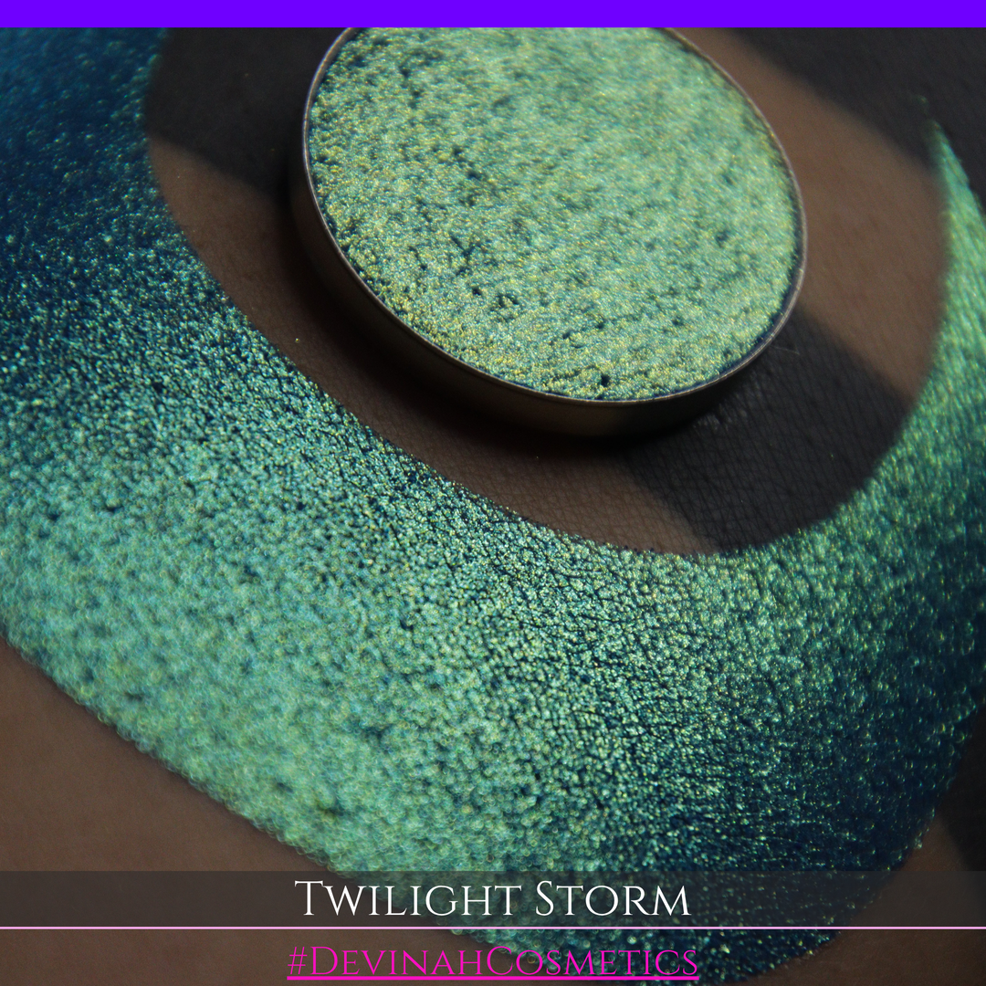 A close-up of a person's arm with a large crescent-shaped eyeshadow swatch in a shimmering teal-to-lime green gradient, with a round pan of the same eyeshadow placed above it. Text at the bottom reads 'TWILIGHT STORM' and '#DEVINAHCOSMETICS'.
