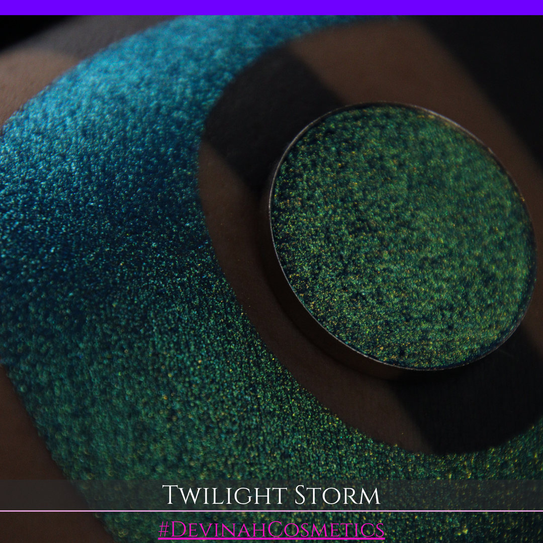 A close-up of a person's arm with a large crescent-shaped eyeshadow swatch in a shimmering teal-to-lime green gradient, with a round pan of the same eyeshadow placed above it. Text at the bottom reads 'TWILIGHT STORM' and '#DEVINAHCOSMETICS'.