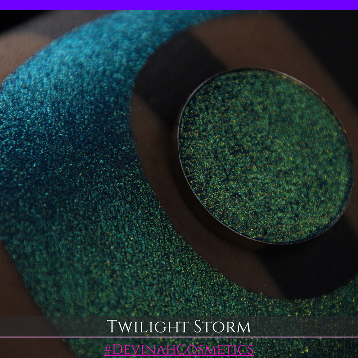 A close-up of a person's arm with a large crescent-shaped eyeshadow swatch in a shimmering teal-to-lime green gradient, with a round pan of the same eyeshadow placed above it. Text at the bottom reads 'TWILIGHT STORM' and '#DEVINAHCOSMETICS'.