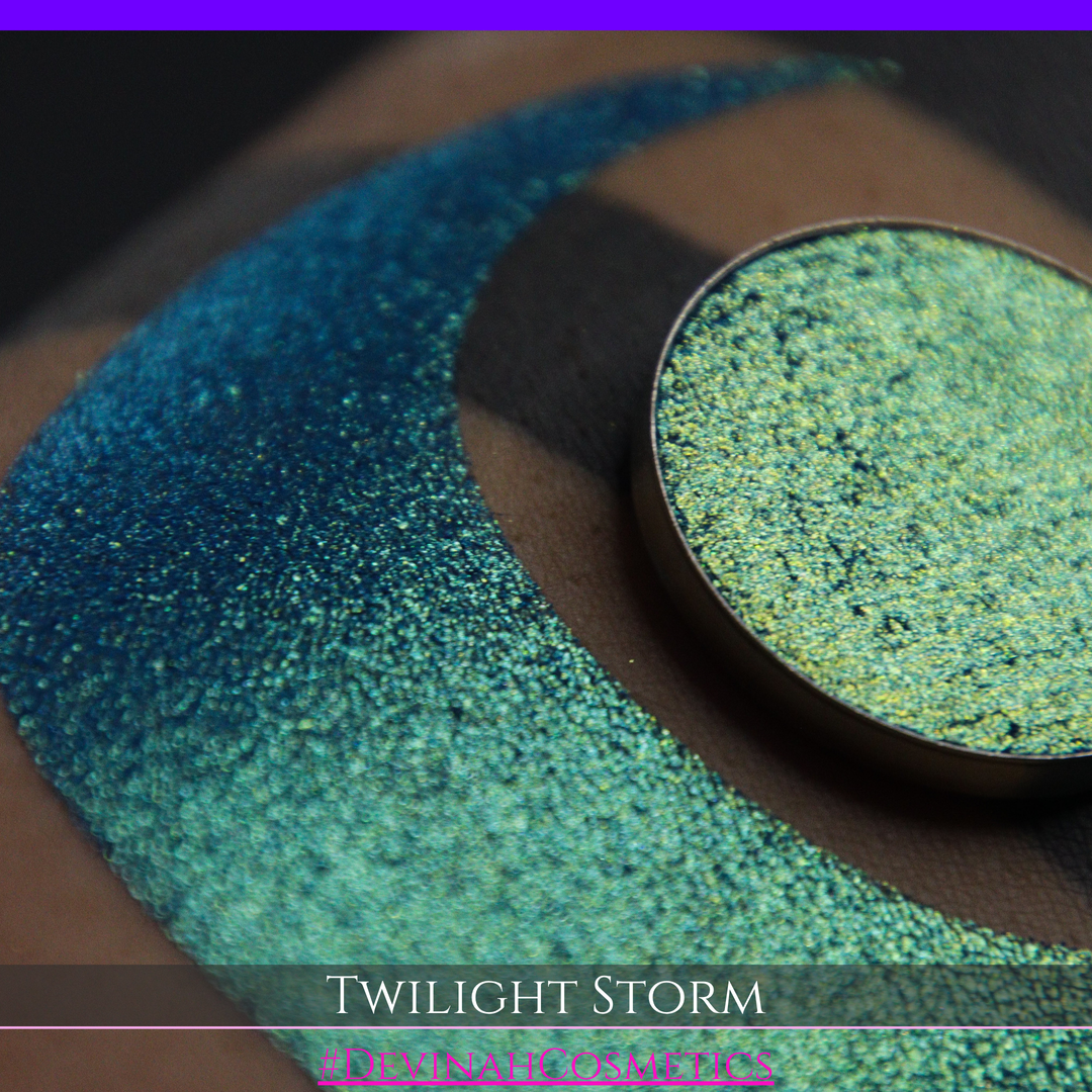 A close-up of a person's arm with a large crescent-shaped eyeshadow swatch in a shimmering teal-to-lime green gradient, with a round pan of the same eyeshadow placed above it. Text at the bottom reads 'TWILIGHT STORM' and '#DEVINAHCOSMETICS'.