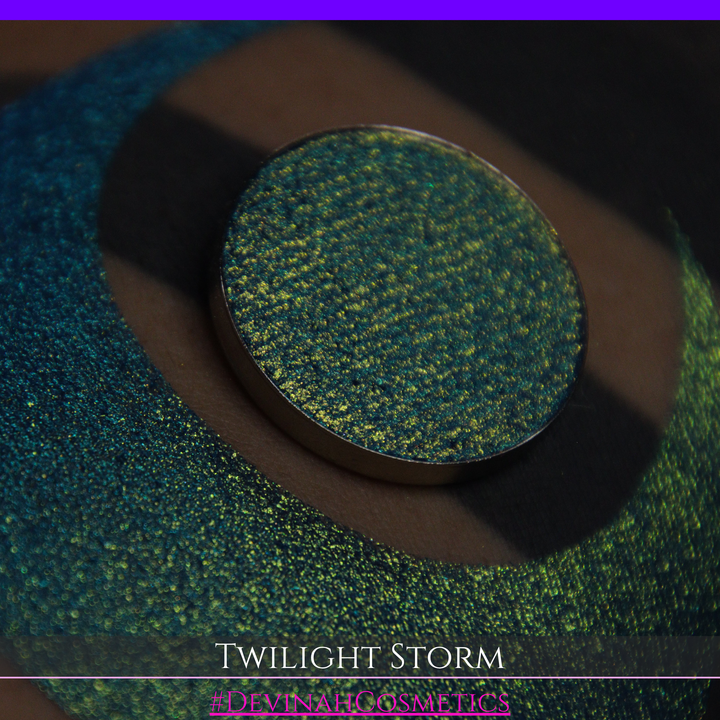 A close-up of a person's arm with a large crescent-shaped eyeshadow swatch in a shimmering teal-to-lime green gradient, with a round pan of the same eyeshadow placed above it. Text at the bottom reads 'TWILIGHT STORM' and '#DEVINAHCOSMETICS'.