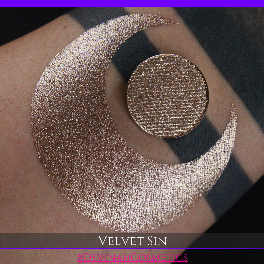 A round eyeshadow pan labeled 'VELVET SIN' made by Devinah Cosmetics, placed over a large crescent moon swatch of the same shimmering shifty eyeshadow on skin. The eyeshadow has colors of beige tan creme colored shimmer.