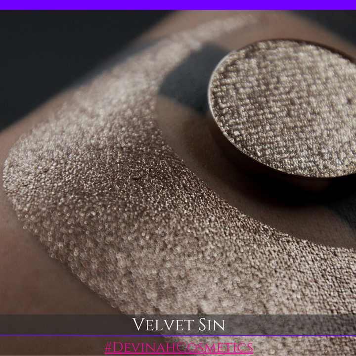 A round eyeshadow pan labeled 'VELVET SIN' made by Devinah Cosmetics, placed over a large crescent moon swatch of the same shimmering shifty eyeshadow on skin. The eyeshadow has colors of beige tan creme colored shimmer.