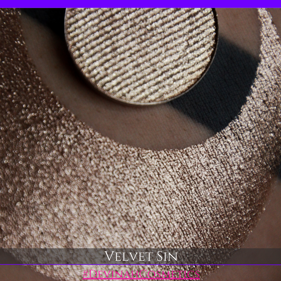 A round eyeshadow pan labeled 'VELVET SIN' made by Devinah Cosmetics, placed over a large crescent moon swatch of the same shimmering shifty eyeshadow on skin. The eyeshadow has colors of beige tan creme colored shimmer.