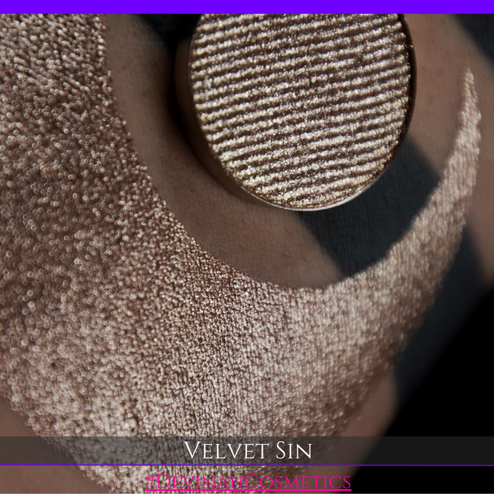 A round eyeshadow pan labeled 'VELVET SIN' made by Devinah Cosmetics, placed over a large crescent moon swatch of the same shimmering shifty eyeshadow on skin. The eyeshadow has colors of beige tan creme colored shimmer.