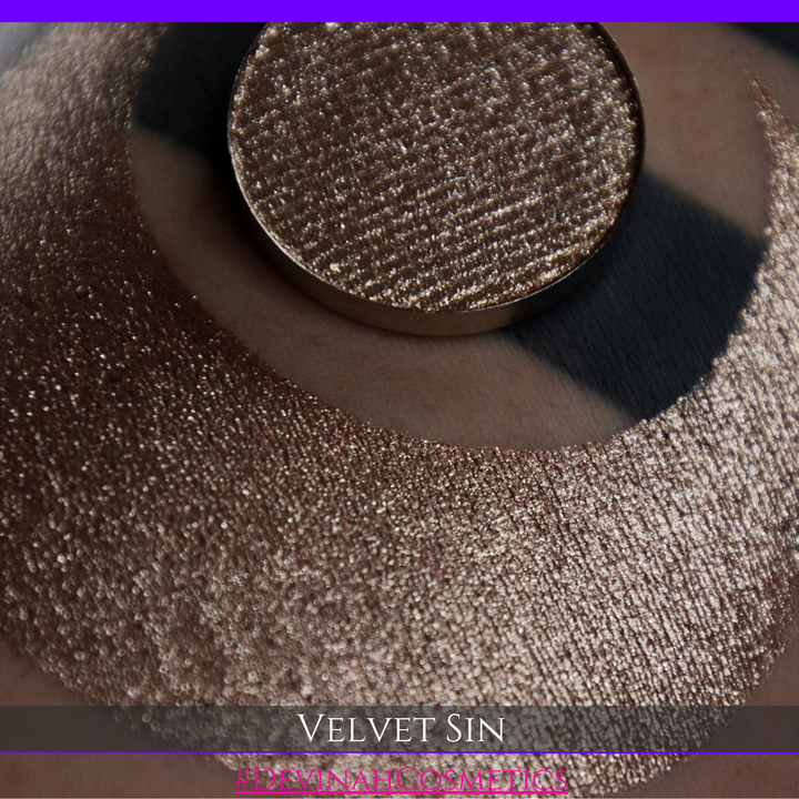 A round eyeshadow pan labeled 'VELVET SIN' made by Devinah Cosmetics, placed over a large crescent moon swatch of the same shimmering shifty eyeshadow on skin. The eyeshadow has colors of beige tan creme colored shimmer.
