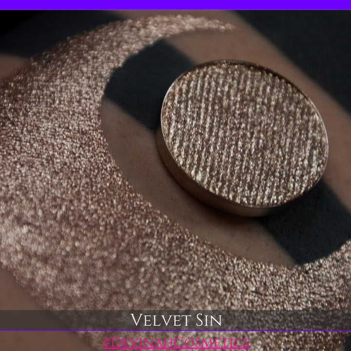 A round eyeshadow pan labeled 'VELVET SIN' made by Devinah Cosmetics, placed over a large crescent moon swatch of the same shimmering shifty eyeshadow on skin. The eyeshadow has colors of beige tan creme colored shimmer.