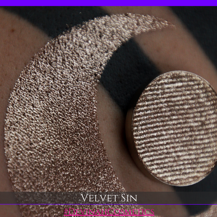 A round eyeshadow pan labeled 'VELVET SIN' made by Devinah Cosmetics, placed over a large crescent moon swatch of the same shimmering shifty eyeshadow on skin. The eyeshadow has colors of beige tan creme colored shimmer.