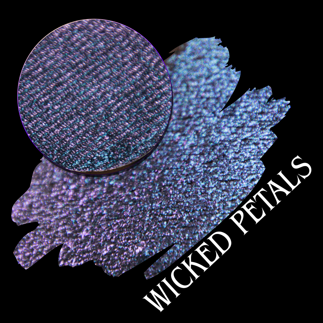 A round eyeshadow pan labeled 'WICKED PETALS' made by Devinah Cosmetics, placed over a large crescent moon swatch of the same shimmering shifty eyeshadow on skin. The eyeshadow has a duochrome multichrome shift between colors blue, purple, pink.