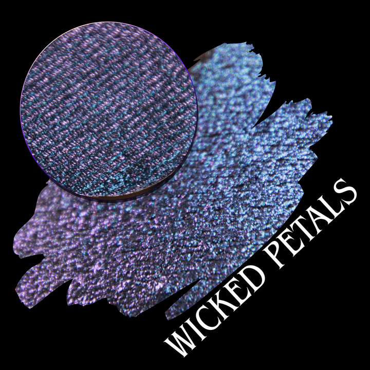 A round eyeshadow pan labeled 'WICKED PETALS' made by Devinah Cosmetics, placed over a large crescent moon swatch of the same shimmering shifty eyeshadow on skin. The eyeshadow has a duochrome multichrome shift between colors blue, purple, pink.