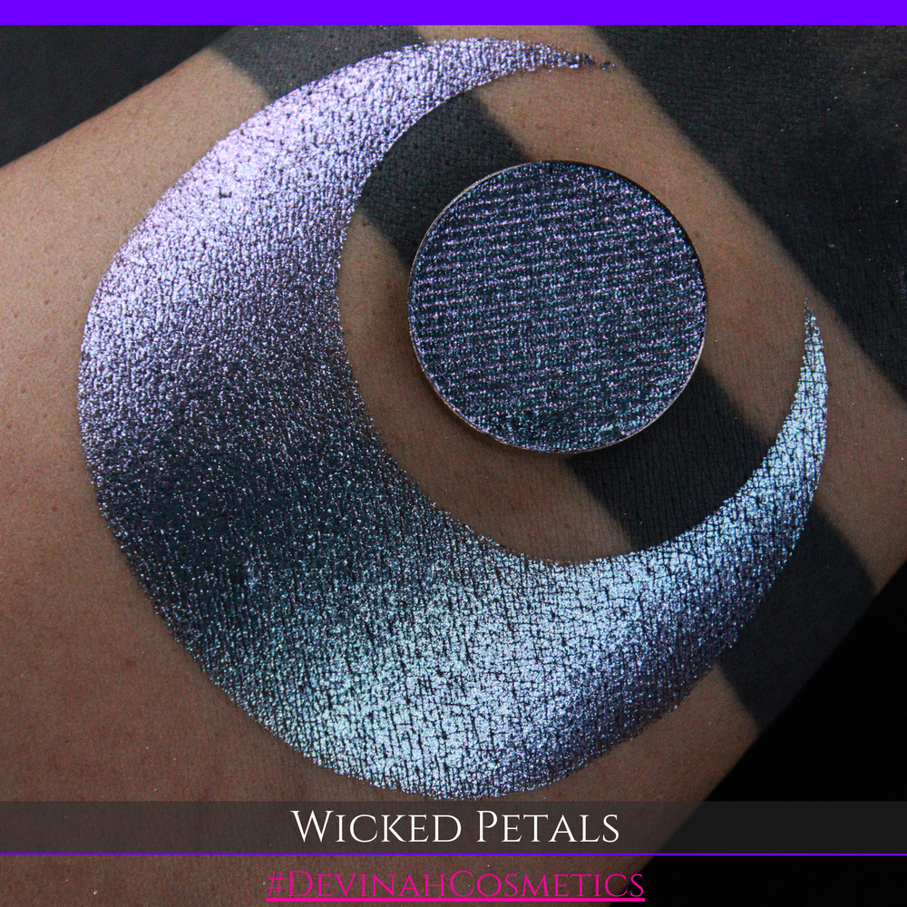 A round eyeshadow pan labeled 'WICKED PETALS' made by Devinah Cosmetics, placed over a large crescent moon swatch of the same shimmering shifty eyeshadow on skin. The eyeshadow has a duochrome multichrome shift between colors blue, purple, pink.