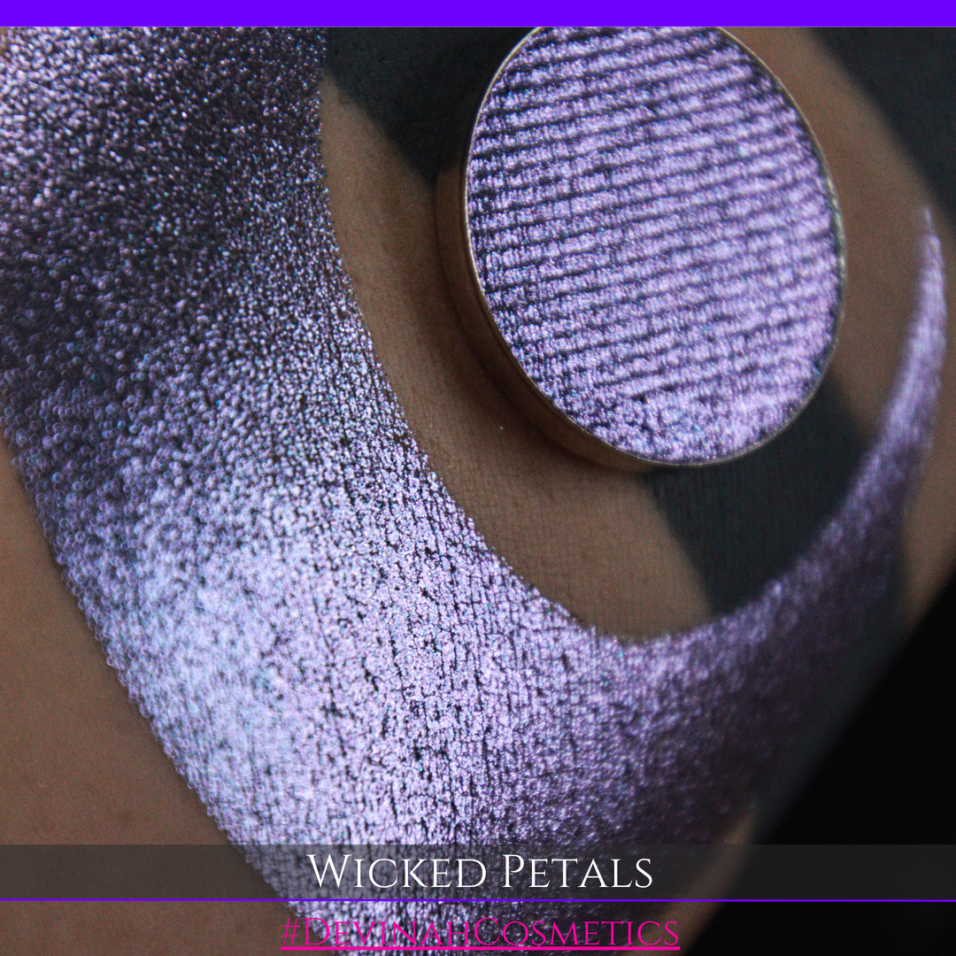 A round eyeshadow pan labeled 'WICKED PETALS' made by Devinah Cosmetics, placed over a large crescent moon swatch of the same shimmering shifty eyeshadow on skin. The eyeshadow has a duochrome multichrome shift between colors blue, purple, pink.