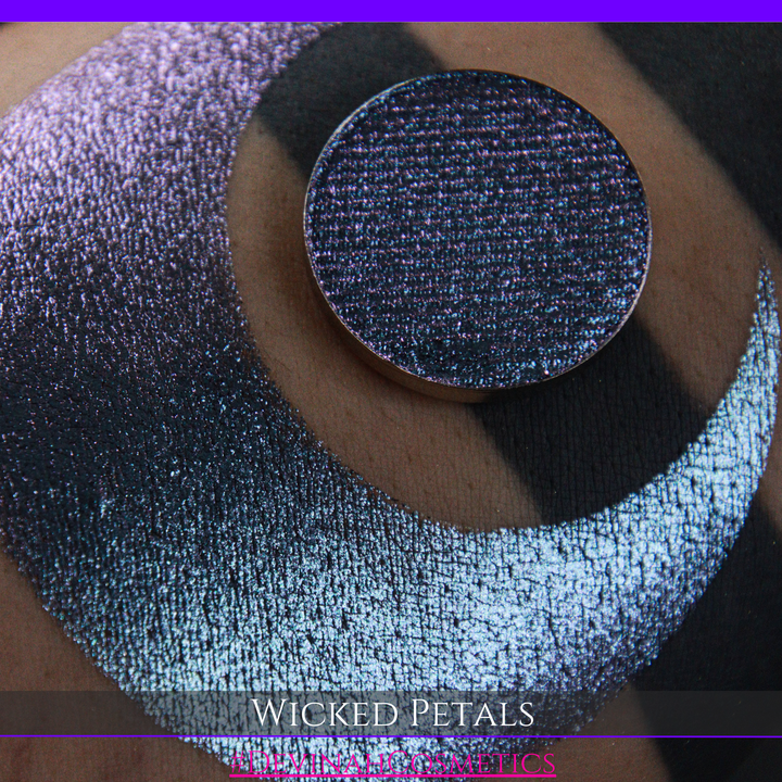 A round eyeshadow pan labeled 'WICKED PETALS' made by Devinah Cosmetics, placed over a large crescent moon swatch of the same shimmering shifty eyeshadow on skin. The eyeshadow has a duochrome multichrome shift between colors blue, purple, pink.