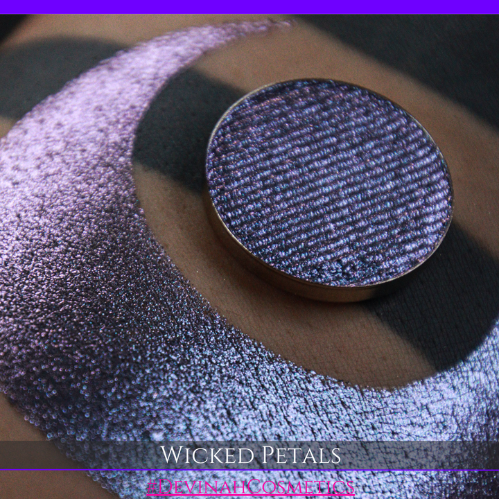 A round eyeshadow pan labeled 'WICKED PETALS' made by Devinah Cosmetics, placed over a large crescent moon swatch of the same shimmering shifty eyeshadow on skin. The eyeshadow has a duochrome multichrome shift between colors blue, purple, pink.