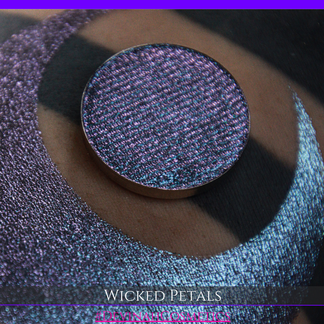 A round eyeshadow pan labeled 'WICKED PETALS' made by Devinah Cosmetics, placed over a large crescent moon swatch of the same shimmering shifty eyeshadow on skin. The eyeshadow has a duochrome multichrome shift between colors blue, purple, pink.