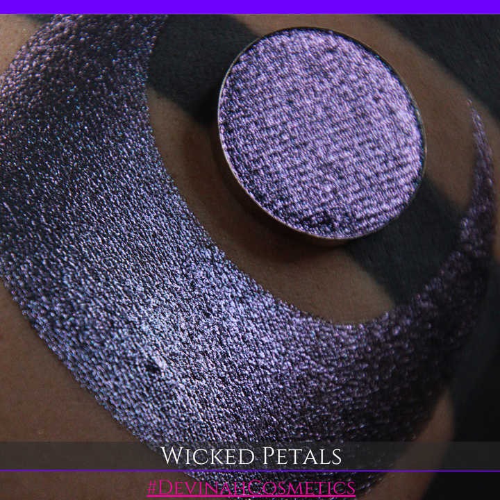 A round eyeshadow pan labeled 'WICKED PETALS' made by Devinah Cosmetics, placed over a large crescent moon swatch of the same shimmering shifty eyeshadow on skin. The eyeshadow has a duochrome multichrome shift between colors blue, purple, pink.