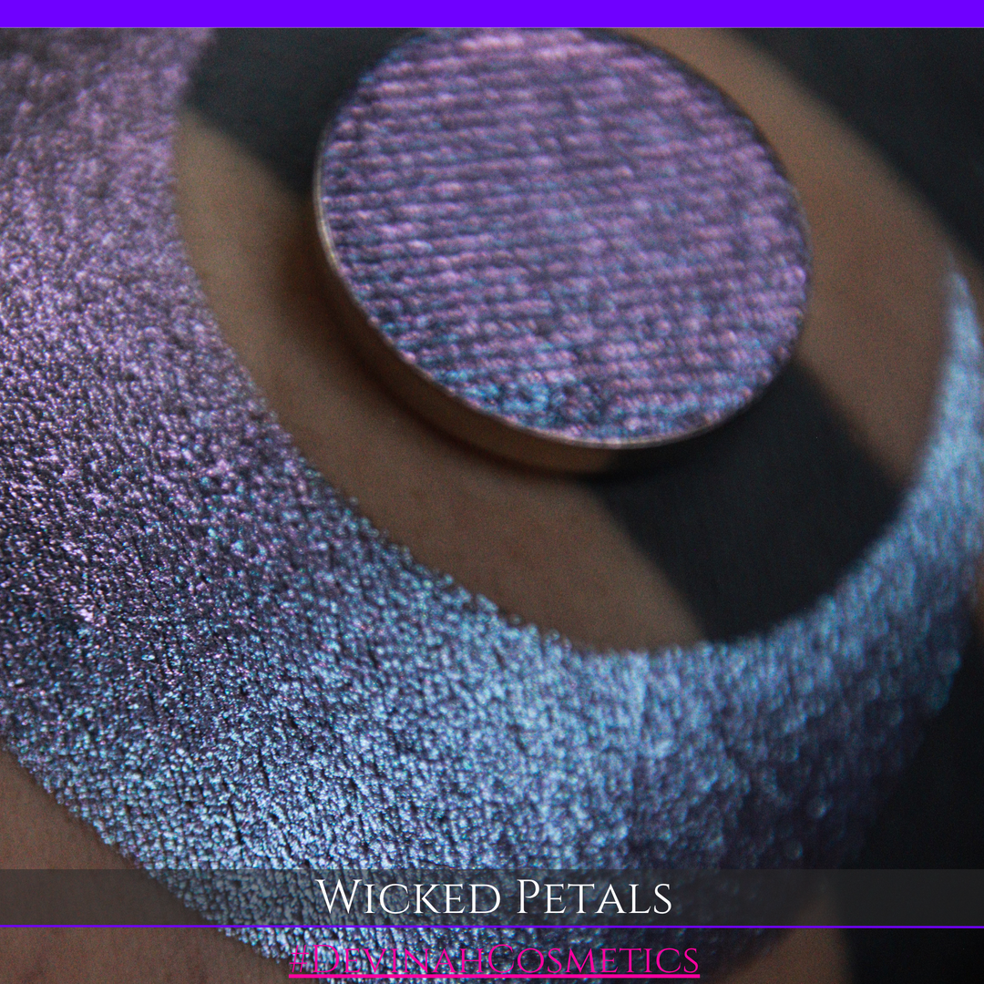 A round eyeshadow pan labeled 'WICKED PETALS' made by Devinah Cosmetics, placed over a large crescent moon swatch of the same shimmering shifty eyeshadow on skin. The eyeshadow has a duochrome multichrome shift between colors blue, purple, pink.