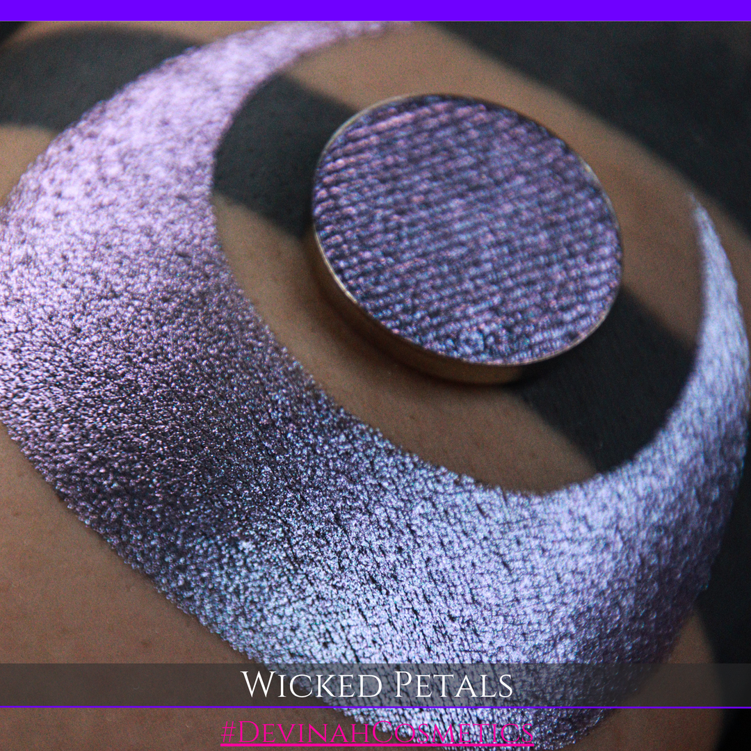 A round eyeshadow pan labeled 'WICKED PETALS' made by Devinah Cosmetics, placed over a large crescent moon swatch of the same shimmering shifty eyeshadow on skin. The eyeshadow has a duochrome multichrome shift between colors blue, purple, pink.
