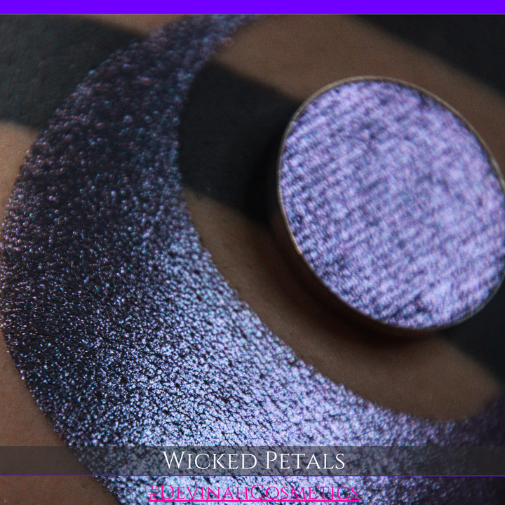 A round eyeshadow pan labeled 'WICKED PETALS' made by Devinah Cosmetics, placed over a large crescent moon swatch of the same shimmering shifty eyeshadow on skin. The eyeshadow has a duochrome multichrome shift between colors blue, purple, pink.
