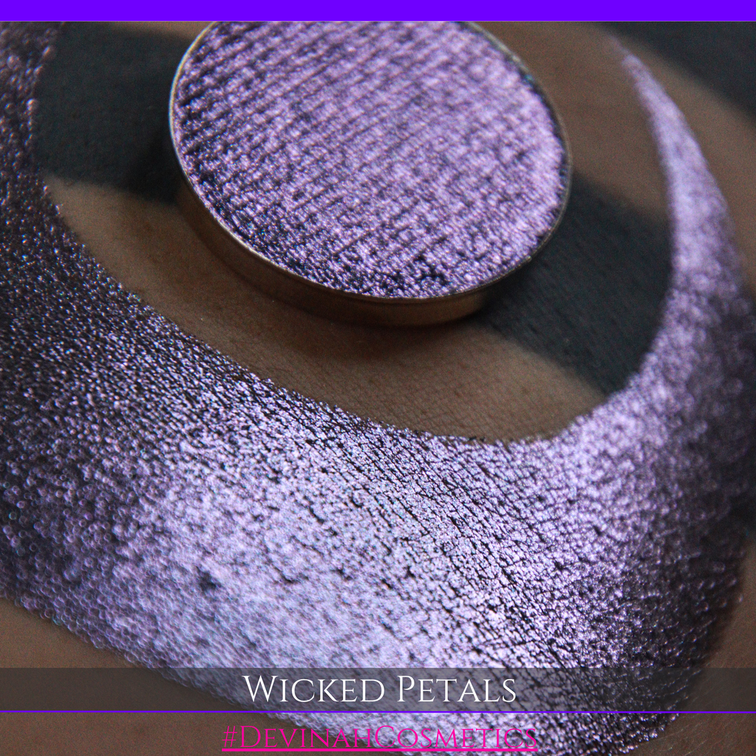 A round eyeshadow pan labeled 'WICKED PETALS' made by Devinah Cosmetics, placed over a large crescent moon swatch of the same shimmering shifty eyeshadow on skin. The eyeshadow has a duochrome multichrome shift between colors blue, purple, pink.