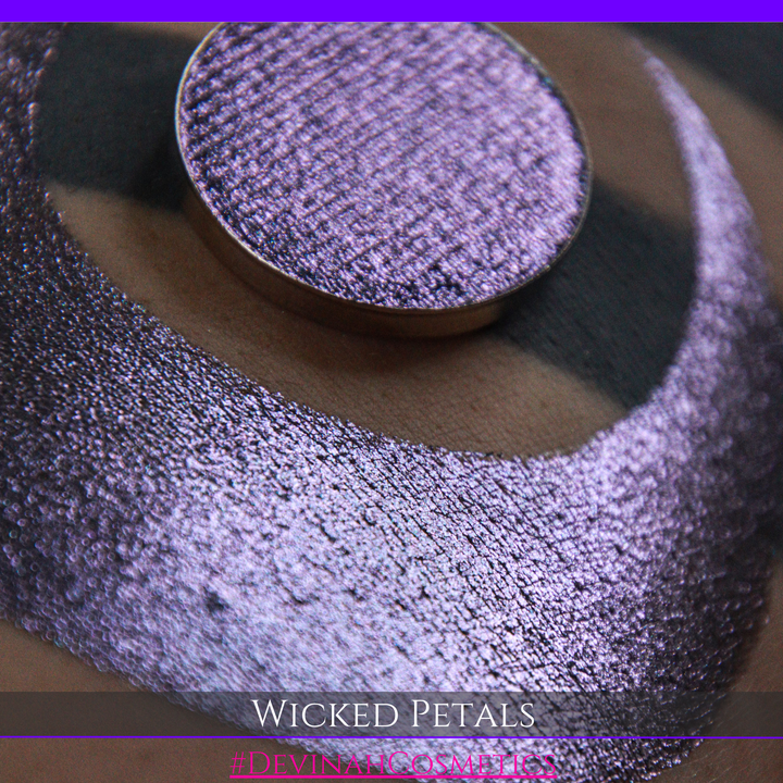 A round eyeshadow pan labeled 'WICKED PETALS' made by Devinah Cosmetics, placed over a large crescent moon swatch of the same shimmering shifty eyeshadow on skin. The eyeshadow has a duochrome multichrome shift between colors blue, purple, pink.