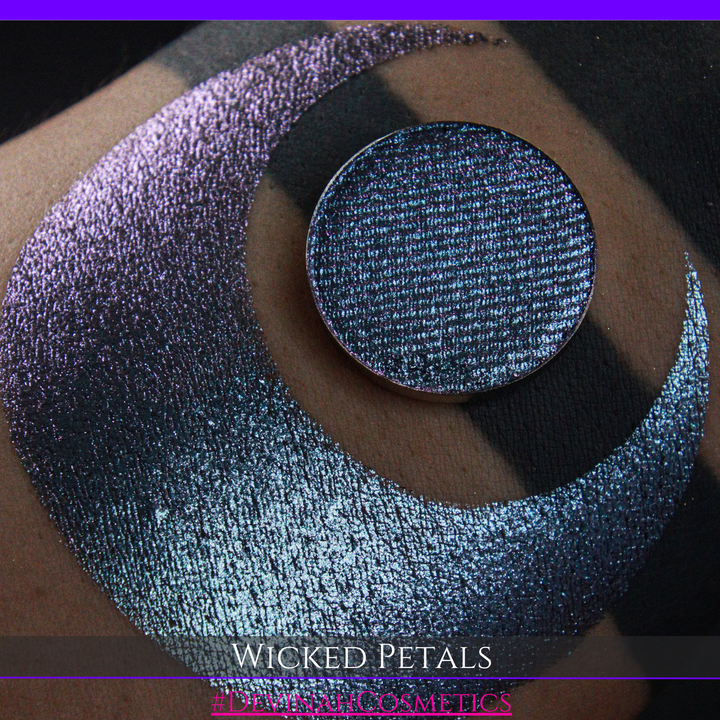 A round eyeshadow pan labeled 'WICKED PETALS' made by Devinah Cosmetics, placed over a large crescent moon swatch of the same shimmering shifty eyeshadow on skin. The eyeshadow has a duochrome multichrome shift between colors blue, purple, pink.