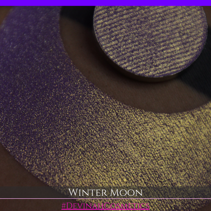 A close-up of a person's arm with a large purple eyeshadow swatch and a matching circular hand made eyeshadow pan, labeled 'Winter Moon' and '#DevinahCosmetics" 