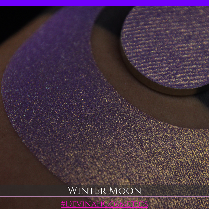 A close-up of a person's arm with a large purple eyeshadow swatch and a matching circular hand made eyeshadow pan, labeled 'Winter Moon' and '#DevinahCosmetics" 