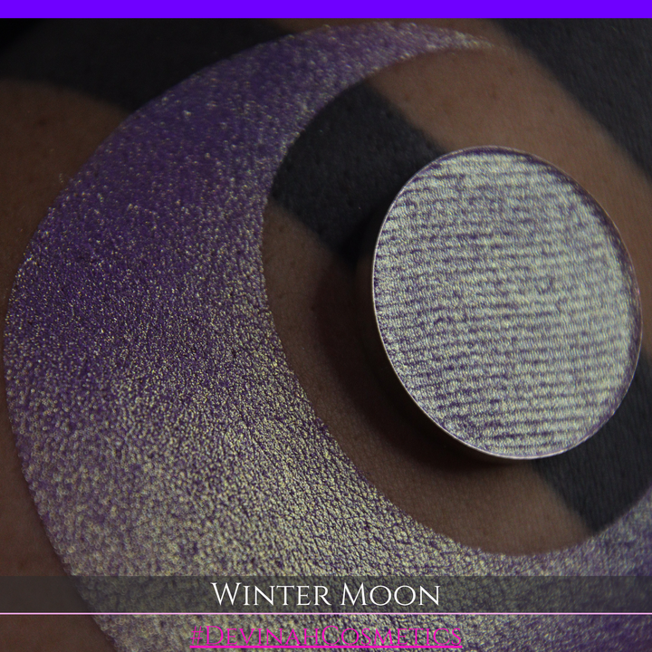 A close-up of a person's arm with a large purple eyeshadow swatch and a matching circular hand made eyeshadow pan, labeled 'Winter Moon' and '#DevinahCosmetics" 
