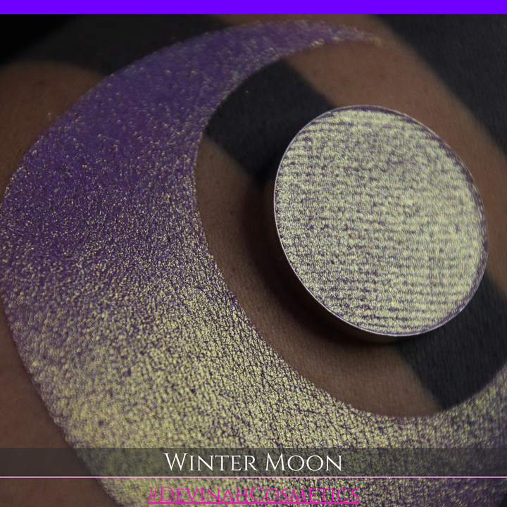 A close-up of a person's arm with a large purple eyeshadow swatch and a matching circular hand made eyeshadow pan, labeled 'Winter Moon' and '#DevinahCosmetics" 
