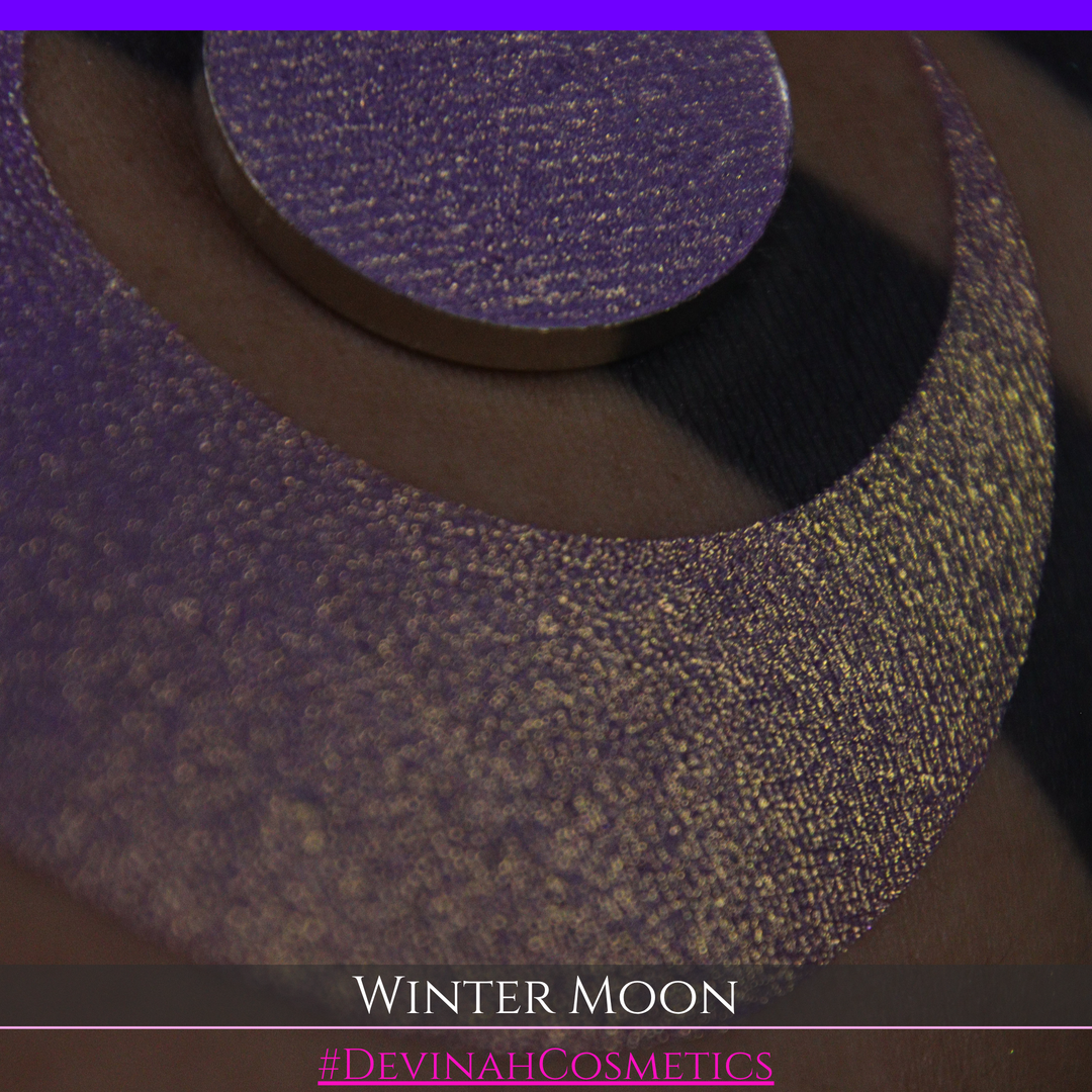 A close-up of a person's arm with a large purple eyeshadow swatch and a matching circular hand made eyeshadow pan, labeled 'Winter Moon' and '#DevinahCosmetics" 